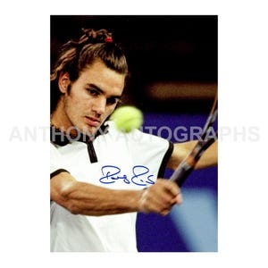May include: A photograph of a tennis player in a white shirt with black trim, holding a racket and hitting a yellow tennis ball. The image includes a signature in blue ink. The player has dark hair tied back.
