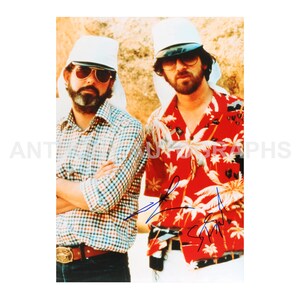 May include: A vintage photograph featuring two men, both wearing sunglasses and white hats. One man is in a plaid shirt, the other in a red floral shirt. Both men have signatures on the image.