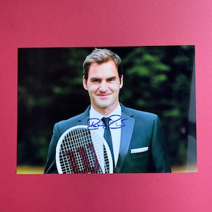 May include: A glossy photograph featuring a man in a black suit holding a tennis racket. The man is smiling and the photo has an autograph in blue ink. The background is a blurred green and the photo is set against a pink background.