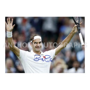 May include: A photograph of a tennis player, wearing a white shirt, headband, and wristband, raising his arms in celebration. He holds a tennis racket in one hand and has a signature on his shirt. The background is blurred with spectators.