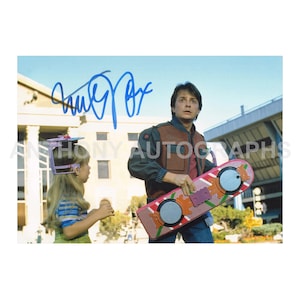 Puede incluir: Fotografía firmada con Michael J. Fox de la película "Regreso al Futuro". Fox sostiene un aeropatín rosa y rojo. Una niña con un tocado también está en la imagen. El fondo incluye un edificio y cielo azul.
