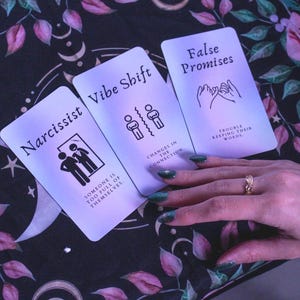 May include: Three tarot cards with the words "Narcissist", "Vibe Shift", and "False Promises". The cards are on a floral patterned cloth. The "Narcissist" card has an image of two people. The "Vibe Shift" card has an image of two figures separated by a line.