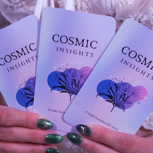 May include: Three Cosmic Insights cards are held in a person's hands. The cards have a lavender background with the words "COSMIC INSIGHTS" in black. Each card features a blue and purple floral design. The copyright reads "Lady Jazmine LLC 2023".
