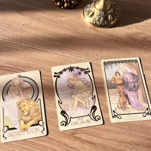 May include: Three tarot cards with Art Nouveau-style illustrations are displayed on a wooden surface. The cards feature images of a woman with a lion, a woman under stars, and figures with a dog. The cards are labeled "Strength", "The Star", and "Ten of Pentacles".