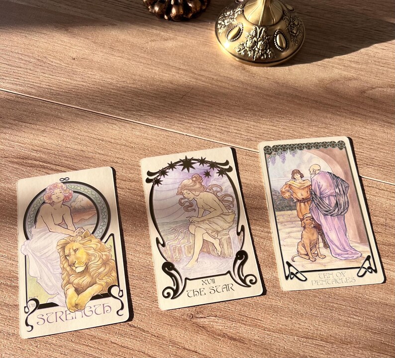 May include: Three tarot cards with Art Nouveau-style illustrations are displayed on a wooden surface. The cards feature images of a woman with a lion, a woman under stars, and figures with a dog. The cards are labeled "Strength", "The Star", and "Ten of Pentacles".