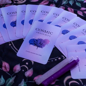 May include: A collection of Cosmic Insights oracle cards with a blue and purple floral design, arranged on a patterned cloth. Two candles, one white and one purple, are placed near the cards. The cards have the text "COSMIC INSIGHTS".