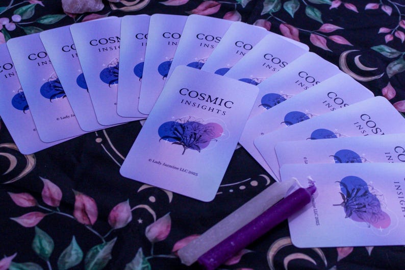 May include: A collection of Cosmic Insights oracle cards with a blue and purple floral design, arranged on a patterned cloth. Two candles, one white and one purple, are placed near the cards. The cards have the text "COSMIC INSIGHTS".