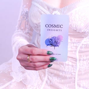 May include: A rectangular card with the text "COSMIC INSIGHTS" and a floral design in blue, purple, and pink is held up. The card is held in front of a white lace dress. The person holding the card has green nail polish and a gold ring.