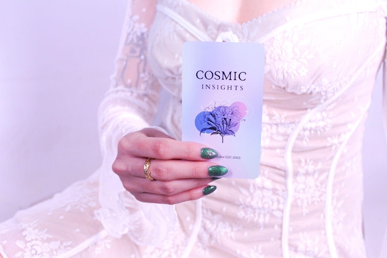 May include: A rectangular card with the text "COSMIC INSIGHTS" and a floral design in blue, purple, and pink is held up. The card is held in front of a white lace dress. The person holding the card has green nail polish and a gold ring.