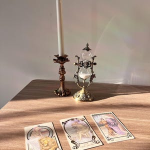 May include: An antique brass candle holder with a tall white candle, a decorative hourglass, and three tarot cards. The cards feature illustrations and the words "Strength" and "The Star". The items are arranged on a wooden surface.