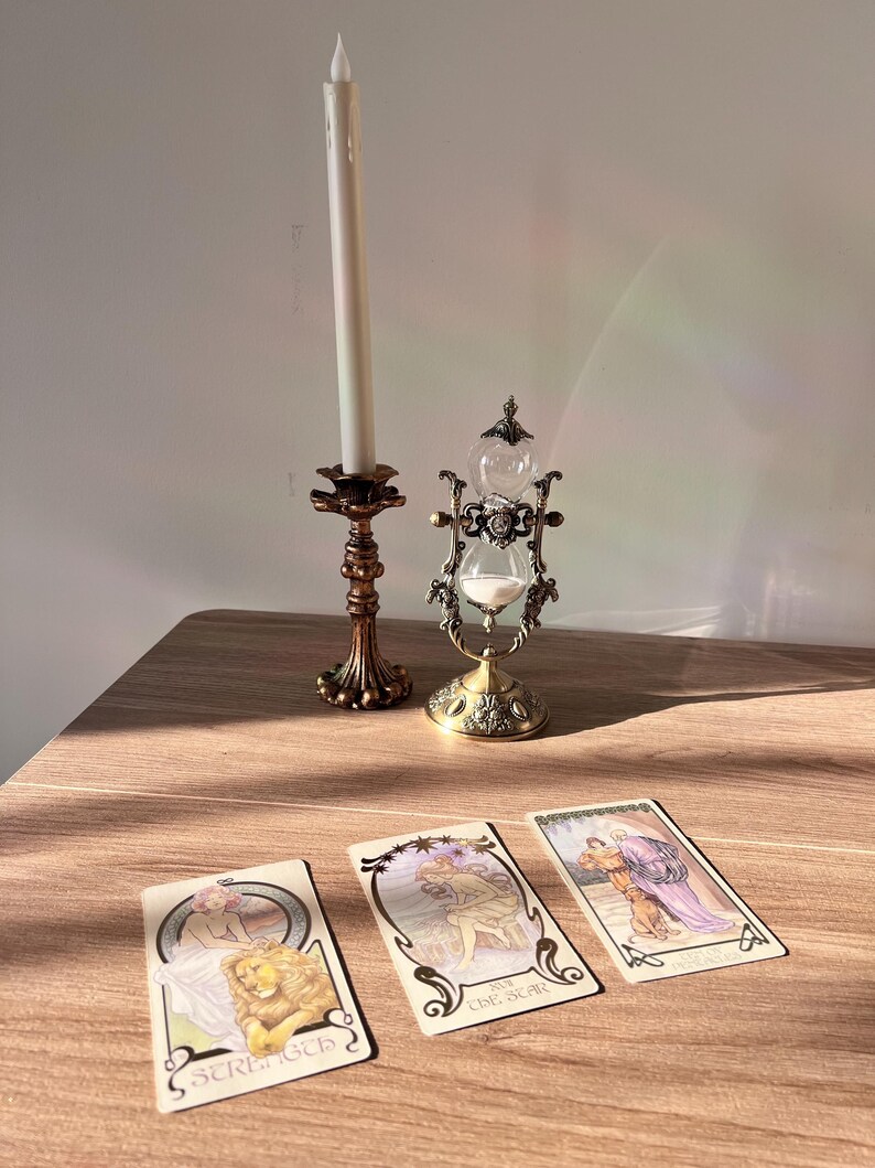 May include: An antique brass candle holder with a tall white candle, a decorative hourglass, and three tarot cards. The cards feature illustrations and the words "Strength" and "The Star". The items are arranged on a wooden surface.