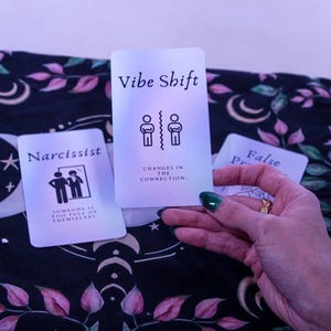 May include: Three tarot cards with text and illustrations are displayed. The center card reads "Vibe Shift" with a graphic of two figures separated by a line. The left card says "Narcissist" with a graphic of two figures. The cards are on a floral patterned cloth.