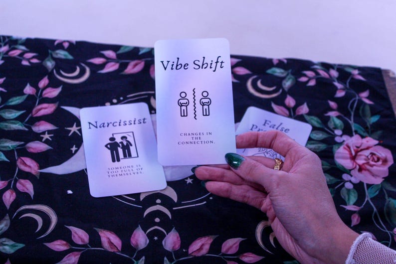 May include: Three tarot cards with text and illustrations are displayed. The center card reads "Vibe Shift" with a graphic of two figures separated by a line. The left card says "Narcissist" with a graphic of two figures. The cards are on a floral patterned cloth.