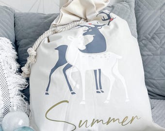 May include: A white canvas gift bag with a drawstring closure. The bag features a design of three stylised deer in shades of grey and white. The text "Summer" is printed in gold at the bottom of the bag.