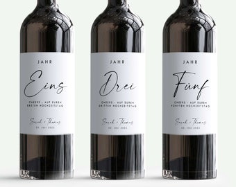 May include: Three wine bottles with white labels. The labels have black text that reads "JAHR" and a number, "Eins", "Drei", or "Fünf". The text below the number reads "CHEERS - AUF EUREN" followed by the anniversary year, "ERSTEN HOCHZEITSTAG", "DRITTEN HOCHZEITSTAG", or "FÜNFTEN HOCHZEITSTAG". The text at the bottom of the label reads "Sarah + Thomas" and "22. JULI 2023".