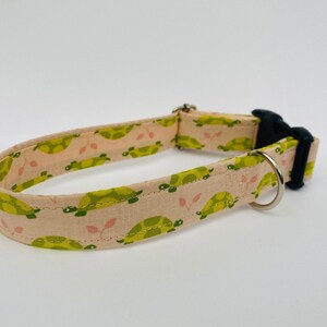 May include: A pink dog collar with a green turtle pattern. The collar has a black buckle and a silver D-ring.
