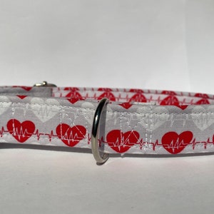 May include: White dog collar with a red heart and EKG pattern. The collar has a black buckle and a silver D-ring.