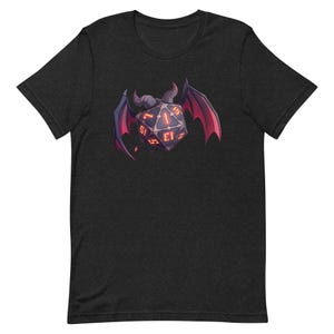 May include: Dark gray t-shirt featuring a graphic of a horned, winged die with glowing red numbers. The die has a dark purple body with red accents on the wings and numbers. A unique design for fans of tabletop gaming.