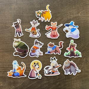May include: A collection of colorful cat stickers with various fantasy-themed designs. The stickers feature cats dressed as wizards, warriors, and musicians, with detailed illustrations and vibrant colors. The stickers are arranged on a dark wooden surface.