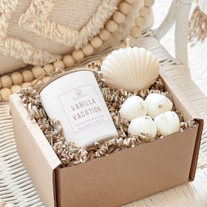 May include: A brown gift box filled with shredded paper, a white candle with the text "Vanilla Vacation" and a seashell-shaped candle. The box is sitting on a white wicker chair with a wooden bead garland.