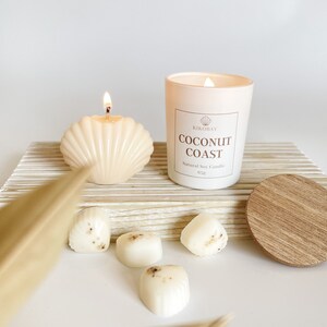 May include: A white soy candle with the text "Coconut Coast" and the brand name "Kikobay" on the label. The candle is surrounded by small white candles and a wooden lid.