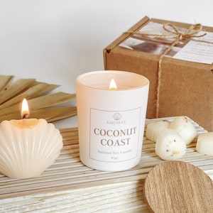 May include: A white candle with a label that reads "KIKOBAY COCONUT COAST Natural Soy Candle 65g". The candle is sitting on a wooden surface with other white candles and a wooden lid.