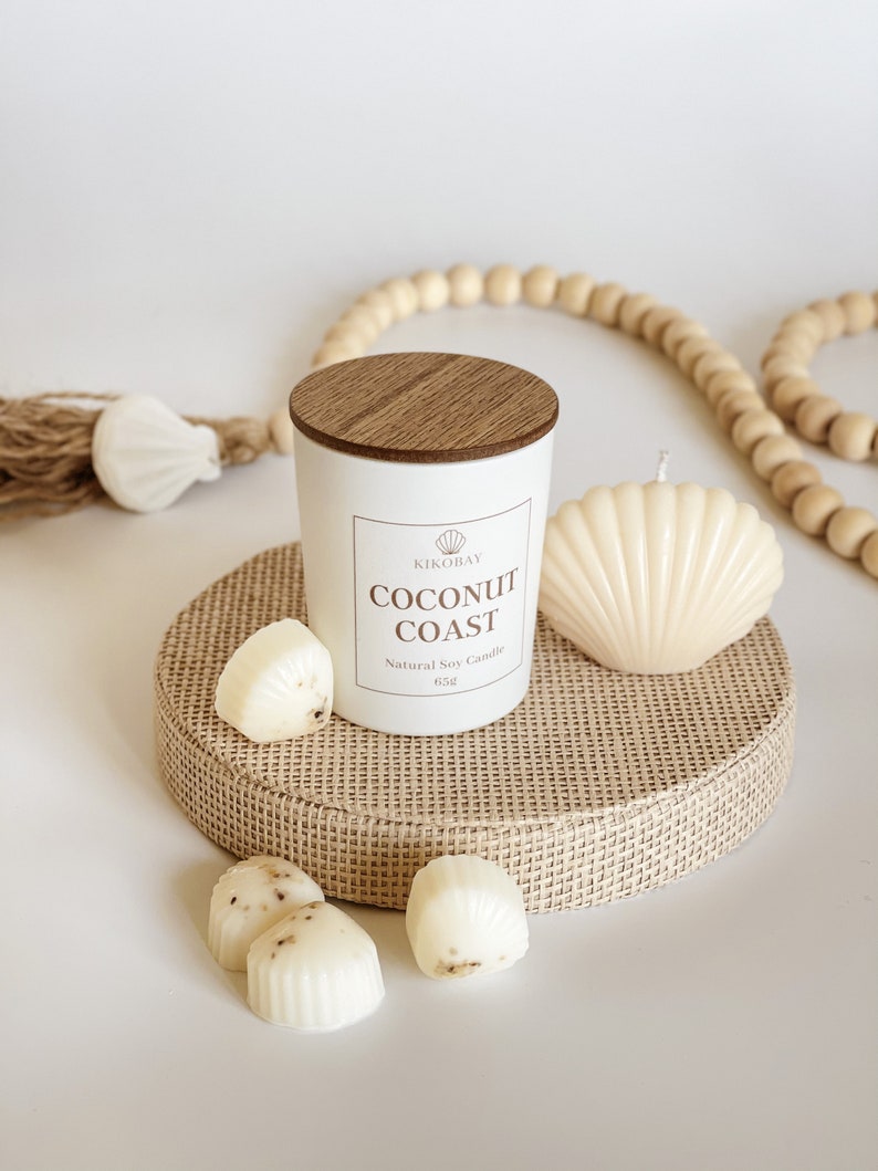 May include: White candle with a wooden lid and a label that reads "Kikobay Coconut Coast Natural Soy Candle 6.5oz". The candle is on a woven mat with a seashell and other small white candles.