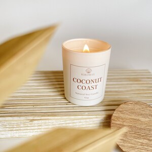 May include: A white candle with a light brown label that reads "KIKOBAY COCONUT COAST Natural Soy Candle 65g". The candle is lit and the flame is visible. The candle is sitting on a wooden surface.