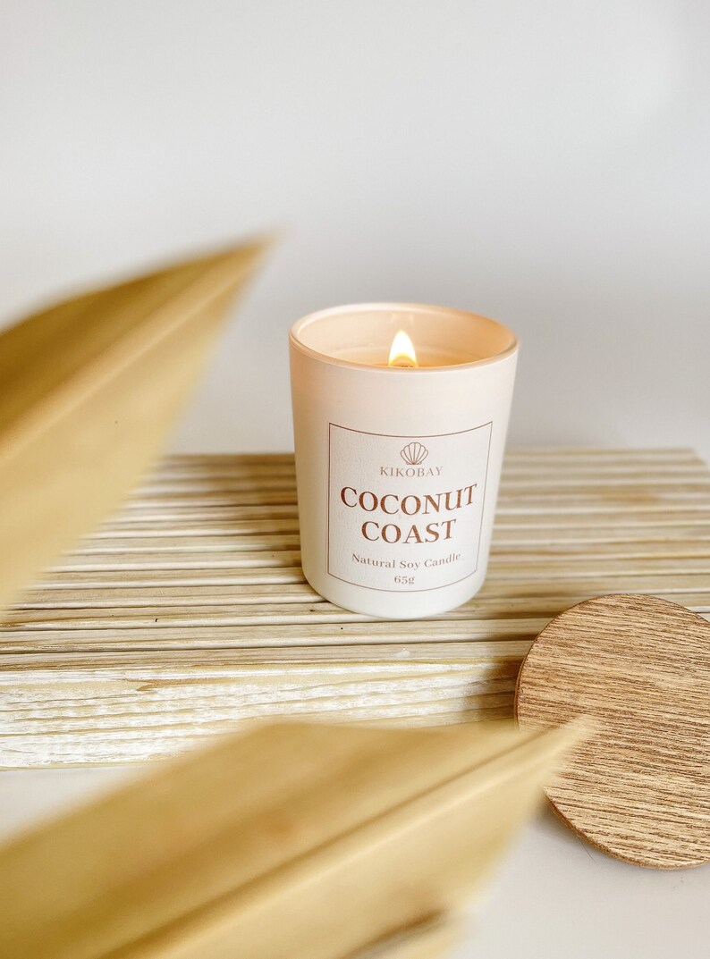 May include: A white candle with a light brown label that reads "KIKOBAY COCONUT COAST Natural Soy Candle 65g". The candle is lit and the flame is visible. The candle is sitting on a wooden surface.