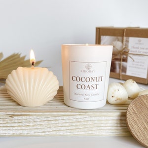 May include: A white soy candle with the text "Coconut Coast" and the brand name "Kikobay" on the label. The candle is sitting on a wooden surface with a seashell candle and a wooden lid.
