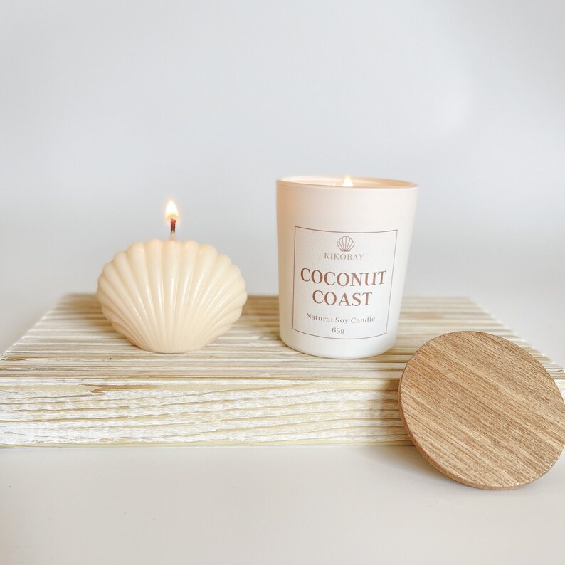 May include: A white candle jar with a wooden lid. The candle is labeled "Coconut Coast" and is a natural soy candle. The candle is sitting on a white wooden surface with a white seashell candle next to it.