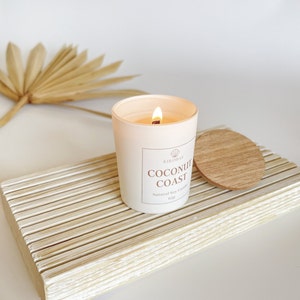May include: A white candle with a wooden lid on a white wooden tray. The candle is labeled "Coconut Coast" and "Natural Soy Candle 6.5oz".