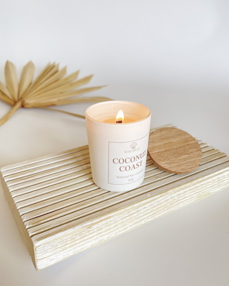 May include: A white candle with a wooden lid on a white wooden tray. The candle is labeled "Coconut Coast" and "Natural Soy Candle 6.5oz".