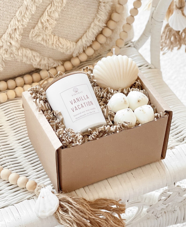 May include: A brown gift box filled with shredded paper, a white candle with the text "Vanilla Vacation" and a seashell-shaped candle. The box is sitting on a white wicker chair with a wooden bead garland.