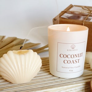 May include: A white candle with a frosted glass jar and a white label that reads "KIKOBAY COCONUT COAST Natural Soy Candle 65g". The candle is lit and there is a plume of smoke rising from the flame. There is a small white seashell candle next to the larger candle.