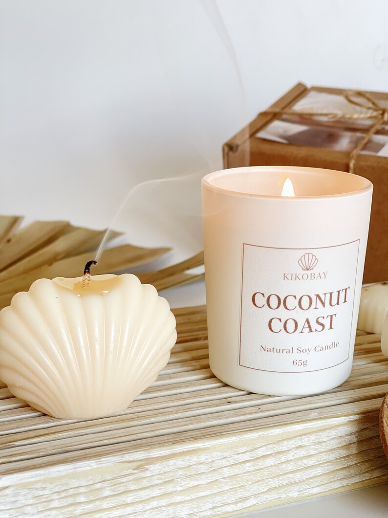 May include: A white candle with a frosted glass jar and a white label that reads "KIKOBAY COCONUT COAST Natural Soy Candle 65g". The candle is lit and there is a plume of smoke rising from the flame. There is a small white seashell candle next to the larger candle.