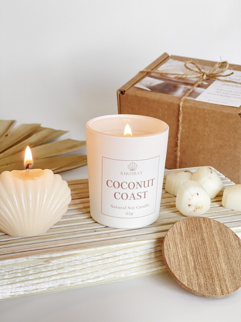 May include: A white candle with a label that reads "KIKOBAY COCONUT COAST Natural Soy Candle 65g". The candle is sitting on a wooden surface with other white candles and a wooden lid.