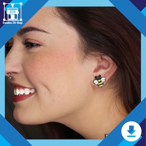 May include: Bee-shaped stud earrings in black, yellow, and white. The earrings are worn on the ear of a person. The earrings have a black outline, yellow body, and white wings. A playful accessory.