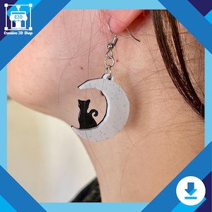 Pode incluir: Brinco prateado com um design de lua crescente e uma silhueta de gato preto. A lua é branca com detalhes salpicados. O brinco é preso a um gancho de peixe prateado.