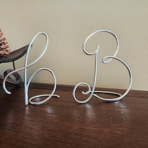 May include: Two silver-colored wire sculptures shaped like the letters b and B. The lowercase b is on the left, and the uppercase B is on the right. Both letters are formed from a single continuous piece of metallic wire with smooth, rounded curves.