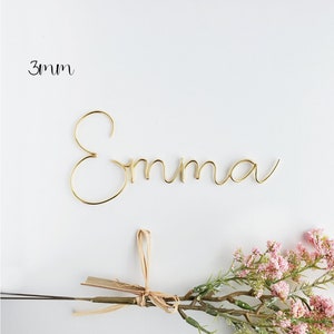 May include: Gold wire name sign spelling "Emma" with a 3mm thickness. The sign is on a white background with a sprig of pink flowers.