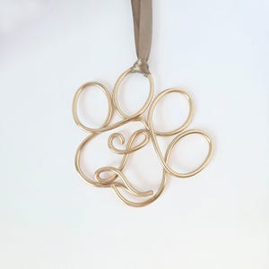 May include: A gold-colored wire ornament in the shape of a paw print, suspended by a taupe ribbon. The paw print design is made of looped wire, creating a delicate and elegant appearance. The ornament is likely for pet lovers.