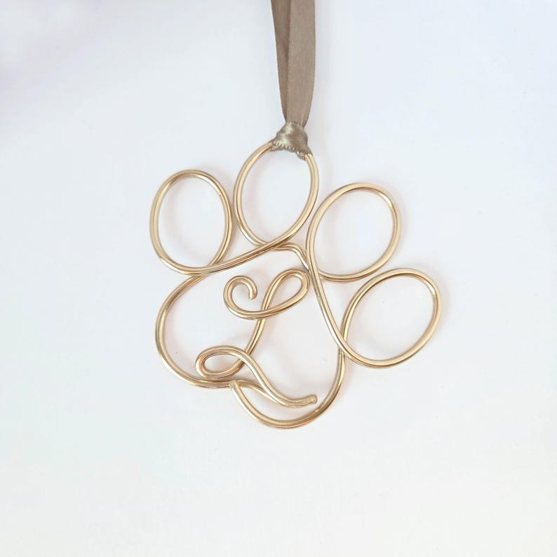 May include: A gold-colored wire ornament in the shape of a paw print, suspended by a taupe ribbon. The paw print design is made of looped wire, creating a delicate and elegant appearance. The ornament is likely for pet lovers.