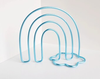 May include: A bright blue metal wire sculpture shaped like a rainbow with four concentric arches. The structure is supported by a base formed from a single continuous wire loop that creates a cloud-like outline at the bottom of the rainbow.