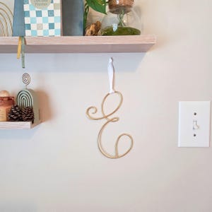 May include: A gold-colored metal wire letter "E" hanging on a wall with a white ribbon. The letter is in a cursive font. The wall is a light beige color. A white light switch is to the right of the letter.