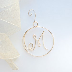 May include: Gold wire ornament shaped like a circle with the letter M in the center. The ornament has a loop at the top for hanging.