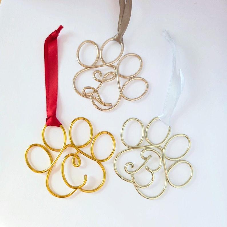 May include: Three wire paw print ornaments in gold, silver, and gold colors, each with a ribbon for hanging. The gold ornament has a red ribbon, the silver has a light blue ribbon, and the other gold ornament has a tan ribbon.