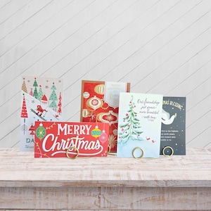 May include: A collection of Christmas greeting cards with various designs, including a red card with "Merry Christmas" text and ornaments, and cards with Christmas trees and winter scenes. Each card is held by a gold-colored metal holder.