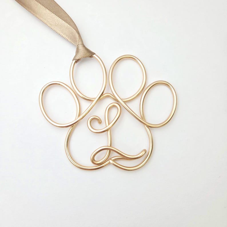 May include: A gold-colored wire paw print ornament with the letter "L" in the center. A gold ribbon is attached at the top for hanging. The ornament is on a white background.