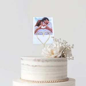 May include: A white tiered cake with a gold heart-shaped photo holder and a photo of a couple. The cake is decorated with a white flower and small white flowers.